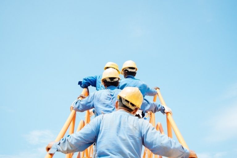 Photo montrant des cadres ouvriers en casque jaunes et en bleu de travail montant les uns derrière les autres une échelle jaune en direction d'un ciel bleu